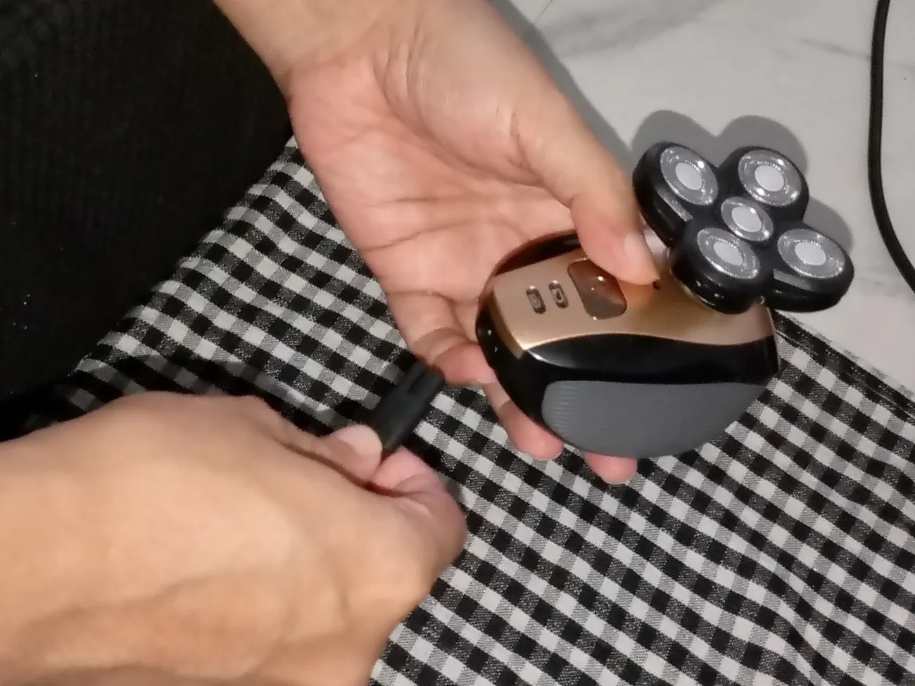 Hands holding and cleaning a gold and black electric shaver with a checkered cloth background.