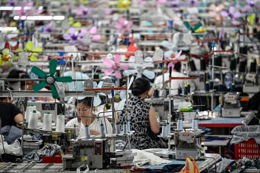 Workers operate sewing machines in a large, crowded textile factory filled with colorful fans and fabric spools.