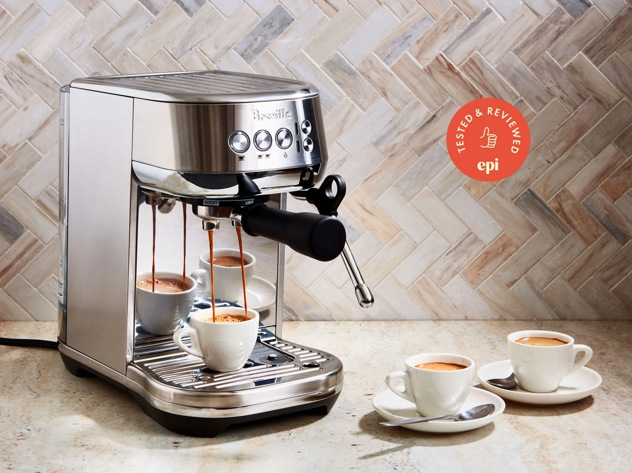 Breville espresso machine brewing coffee into a white cup with two additional cups on saucers nearby on a kitchen counter.