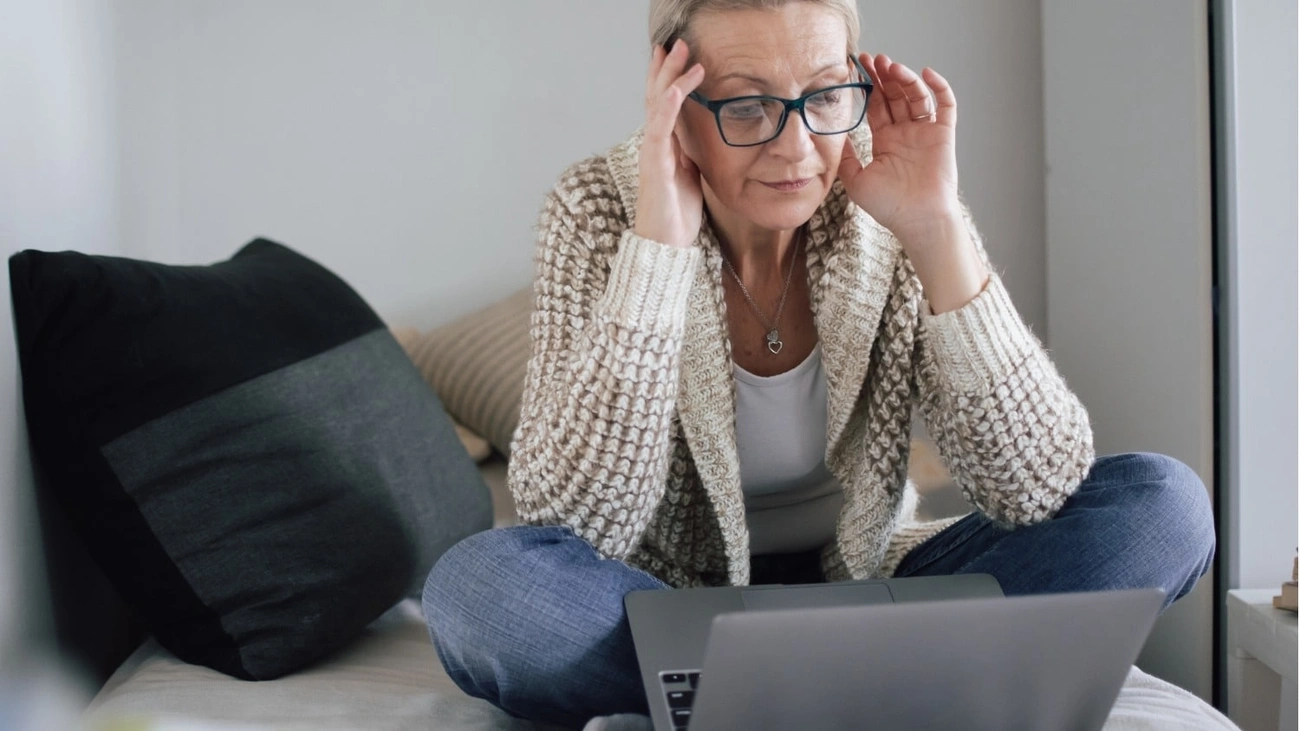 Person wearing a cozy sweater sitting cross-legged on a bed with a laptop, adjusting their glasses.